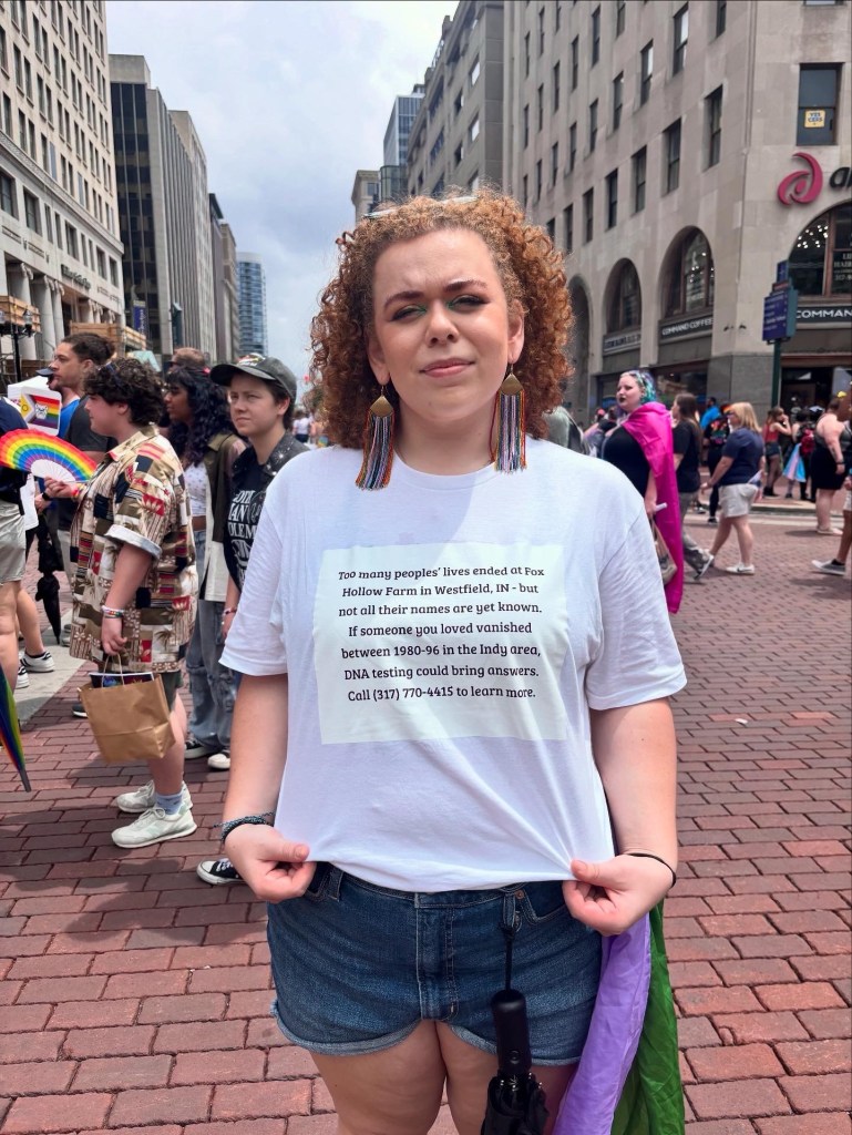 Femme person with curly red hair, white shirt with black lettering, and blue shorts, and brown boots. Letting on the shirt says "Too many peoples' lives ended at Fox Hollow Farm in Westfield IN - but not all their names are yet known. IF someone you loved vanished between 1980-96 in the Indy Area, DNA testing could bring answers. Call (317) 770-4415 to learn more." 