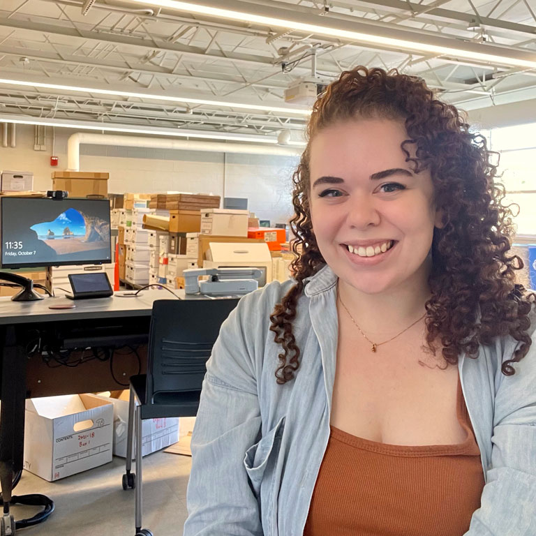 Femme person (Kal Demaree) with brown curly hair, in orange tank top with light blue unbuttoned shirt on top, posed for photo. 