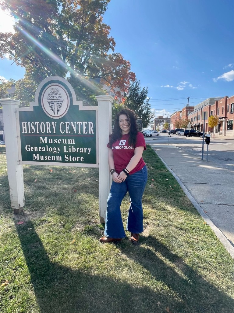 Femme person (Kal Demaree) in red shirt and blue jeans leaning against green and white sign that says "History Center Museum, Genealogy Library, Museum Store"