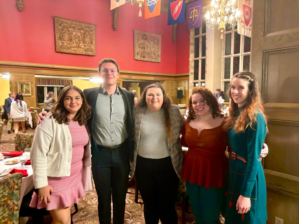 Photo of 5 people. From left to right, a young women with dark hair wearing a pink dress with a white sweater; a tall man wearing a grey shirt and black suit jacket, a women with long brown hair wearing a grey shirt with a brown jacket, a young femme person (Kal Demaree) wearing an orange blouse, and a tall women with brown hair wearing a green dress. The group is posed within the entry way of a banquet hall. 