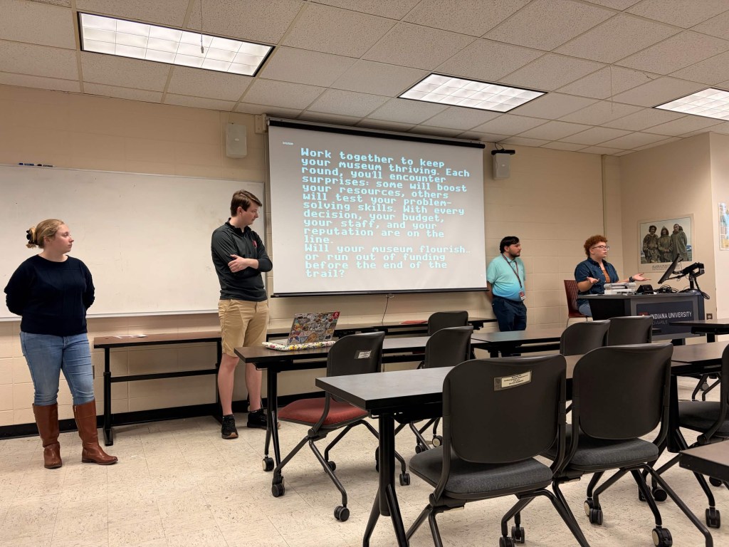 4 people presenting in a classroom in front of a pull down projection screen. On the screen is black background with white lettering. There are desks in front of the students presenting. 