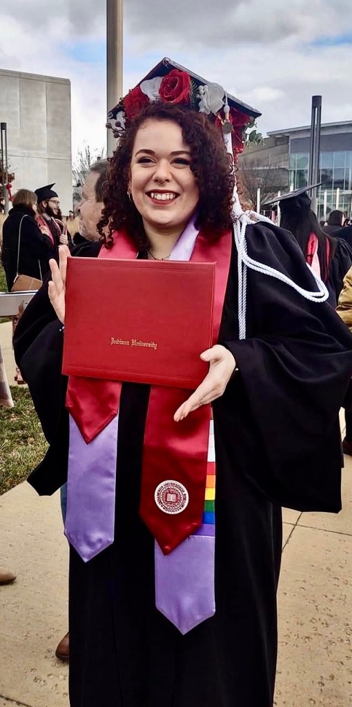 Person (Kal Demaree) with brown curly hair, in black graduation robes and cap (with red and white flowers and ribbons on it), posing for the camera holding red diploma holder. 