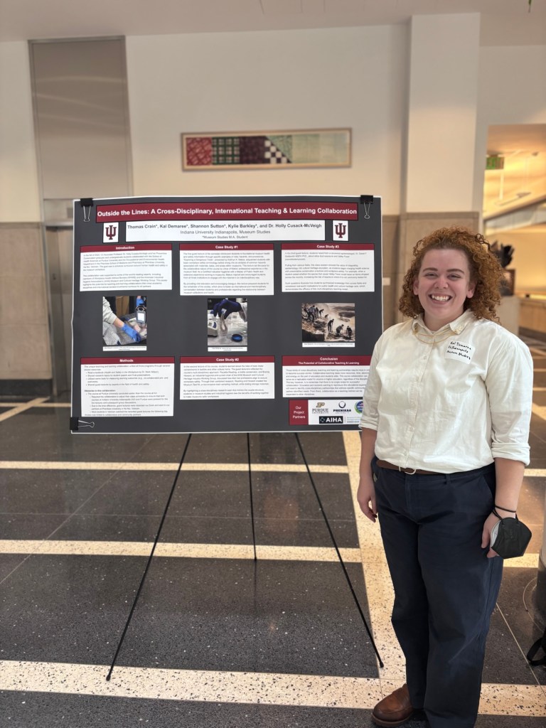 Person (Kal Demaree) with medium legthen curly red/blond hair, in white button down shirt, posed in front of a large black poster with red and white text blocks and images, on an easel.