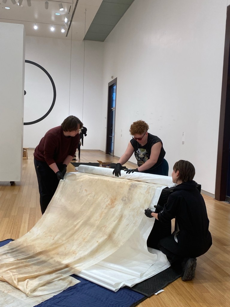 3 people in a white gallery, working together to roll up a long white textile. 