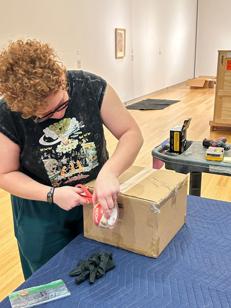 Person with short red curly hair and glasses tapping up a shipping box on a table, within a white walled gallery. 