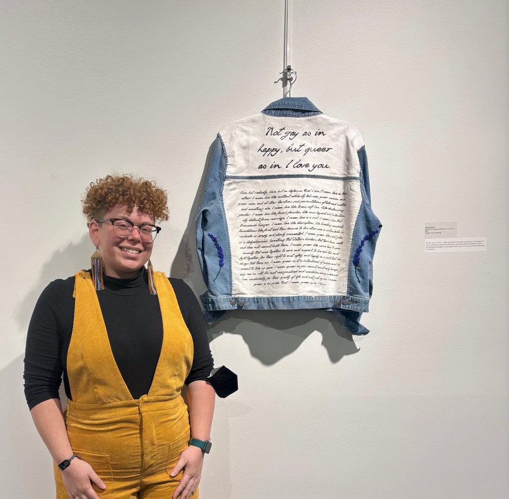 Person (Kal Demaree) is posted to the left of a painted denim jacket. The person has short curly red hair, glasses, and is wearing a black long sleeve shirt with yellow overalls, and rainbow beaded earrings. The blue denim jacket is painted white on the central back panels with black lettering; the lettering at the top says "Not gay as in happy, but queer as in I love you". 