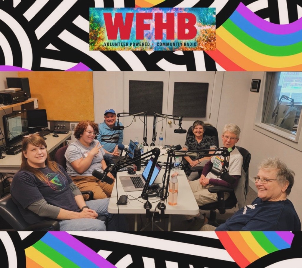 Image of multiple people, in front of a rainbow, black, and white background. A the top of the image is red lettering that says "WFHB Community Radio". In the image of people, there are6 people seated around a table that has black microphones on it.
