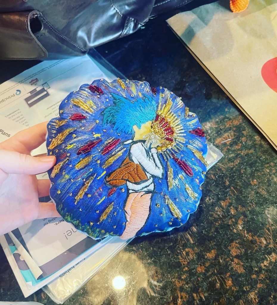 Blue cushion on a black background, on the cushion is an embroidered cartoon image of a person with blue hair swallowing a star. The star is also embroidered, and beaded. 