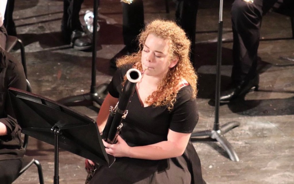 Person (Kal Demaree), with long blond curly hair, in black dress, seated, playing a black bassoon, on a black floored stage as part of a concert band.