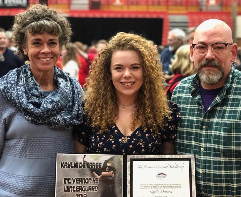 3 people (from left to right, a women with short brown curly hair wearing blue, a shorter younger person with curly blond hair, and a bald man with grey facial hair and glasses. THe middle person is holding a certificate book. 