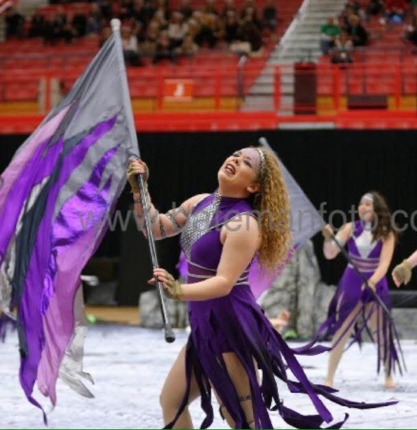 Femme person with long curly red/blond hair, in a purple dress, dancing with a purple and grey flag on a black pole. 
