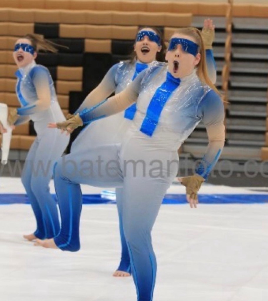 3 people in white and blue leotards with clear plastic vests, and blue plastic masks over their eyes. All three people have long hair in a high pony tail, and are barefoot on a white floor. 