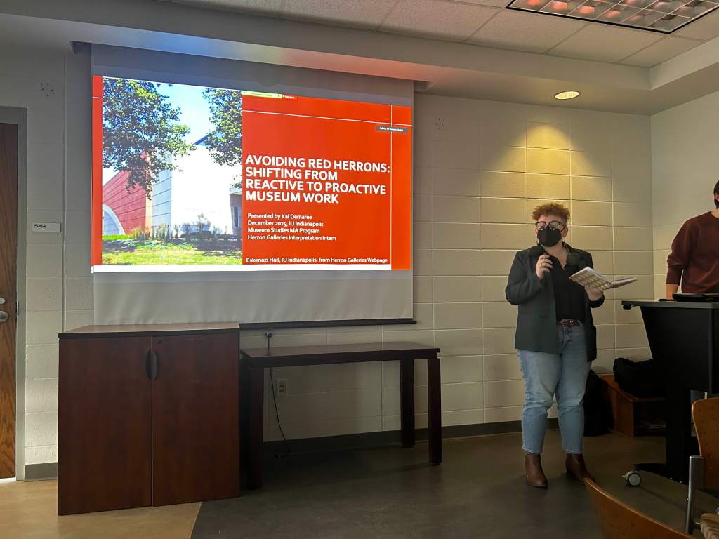 Femme person with short red curly hair in green jacket, in front of a projection screen in a darken room. On the projection screen is a red slide show. 