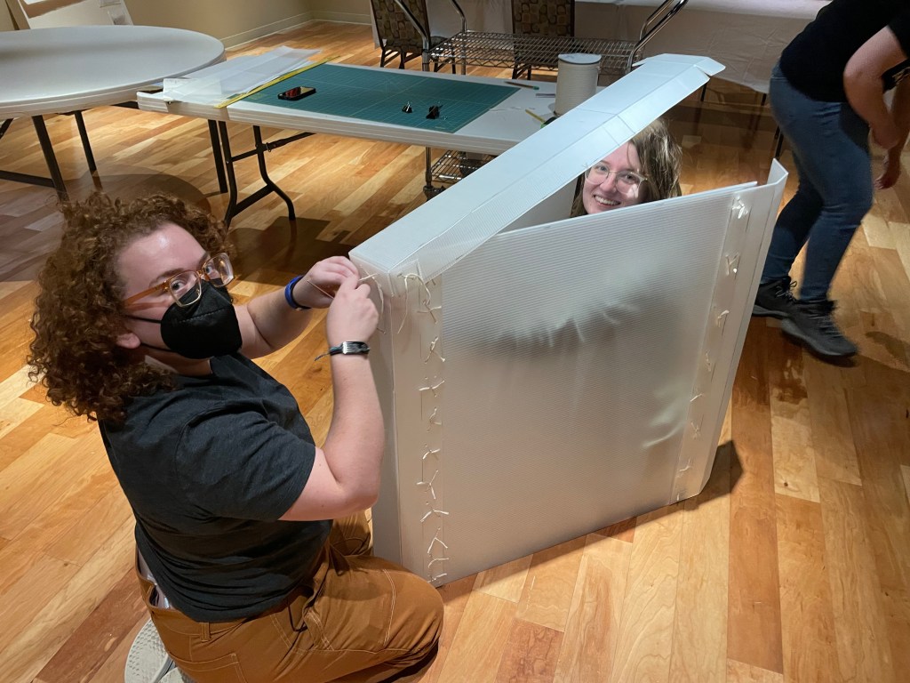 Person (Kal Demaree) with brown curly hair, glasses, and a black N-95 face mask, in black tshirt and brown cargo pants, tying string to attach sections of plastic together to make a box; person with blond hair and glasses is sitting within the open end of the box. 